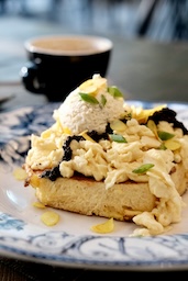 Toast mit Rührei und Schnittlauch, darauf eine weiße Nocke, mit einer Tasse im Hintergrund