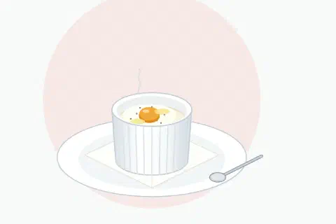 Egg en cocotte with cream in a small white ramekin on a folded napkin, white fully set and a glossy yolk ready to be broken with a spoon